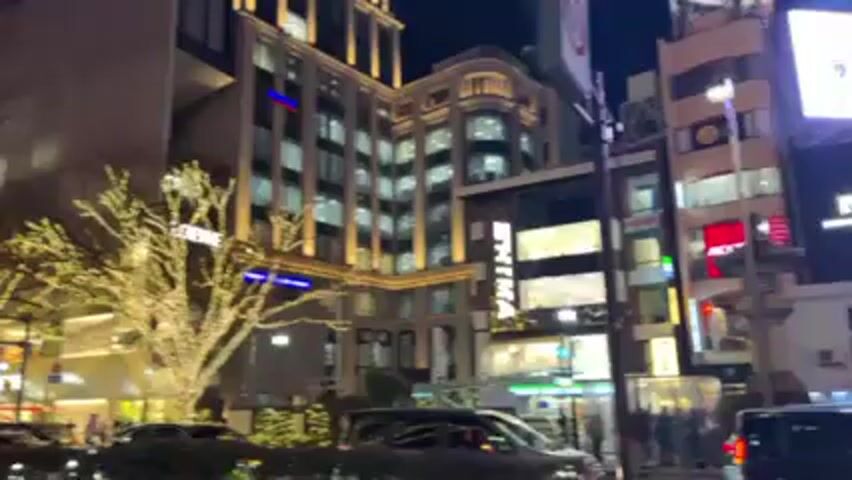 The video shows a nighttime street scene in a city, possibly Japan. A wide street is lined with bare trees covered in white holiday lights. There are several tall buildings on either side of the street, with illuminated windows and commercial signage, including "LOEWE".  Cars and trucks drive along the street, and people can be seen walking on the sidewalks. The overall atmosphere is festive due to the lights.