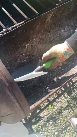 A person is shown in a close-up shot, actively cleaning out a rusty, trough-style barbecue grill outdoors. The individual's arm is visible, covered by a long-sleeved gray and black shirt, and they are wearing a colorful floral-patterned gardening glove on their right hand. They are using a small trowel with a black blade and a bright green handle to scoop gray ashes and small charcoal remnants from the bottom of the grill. With their other hand, they hold open a white plastic bag, into which the collected ashes are being deposited.

The grill itself is old and metallic, showing signs of rust and wear, with a grate visible on top. In the background, on the far side of the grill, a yellow plastic object, possibly a dustpan or another gardening tool, is partially visible. The ground beneath and around the grill is covered with small gray and white stones or gravel. Beyond the immediate area of the grill, green vegetation can be seen, indicating an outdoor, natural setting. The scene is well-lit, suggesting it is daytime with clear weather. The activity takes place in what appears to be a backyard or garden area in Goryachevodsky, Russia. There is no visible text in the video.