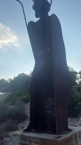 A silhouette statue of a figure wearing a robe and a halo stands against a backdrop of a cloudy sky and the ocean. The statue is dark, appears to be made of metal, and has graffiti on its surface. The figure is holding a long, curved object. The statue is positioned on a rocky outcrop overlooking the water. In the distance, there are some trees and structures. The video is shot from a low angle looking up at the statue.