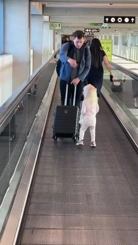 A father and his young daughter are walking on a moving walkway in an airport. The father is wearing a blue jacket and carrying a black rolling suitcase. The daughter, with blonde hair, is wearing a pink shirt and white patterned pants, and holding a white stuffed animal. They are walking towards the camera on the moving walkway, which is made of gray metal slats. In the background, other travelers can be seen, and there are signs with Hebrew text. One sign is green and yellow and appears to be directional, with an arrow and the word "PROTECTED". Another sign has the Hebrew word "ממוגן" which translates to "protected" or "shielded". The overall scene suggests they are navigating through an airport terminal. The lighting indicates it is daytime. The child appears engaged and happy, while the father seems focused on their journey. The setting is a modern airport environment.