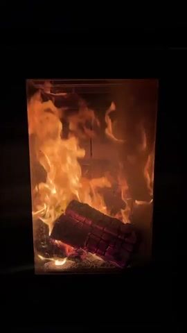 The video shows a fire burning in a rectangular fire pit. There are wooden logs at the base of the fire. The fire pit appears to be constructed of a clear material, possibly glass or plastic, allowing the flames to be seen on all sides. The background is dark, focusing the viewer's attention on the fire.