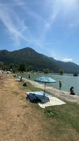 The video shows a sunny day at a crowded beach. In the foreground, a beach umbrella shades a blanket and some towels on the grass. Further back, people are sunbathing and wading in the clear turquoise water. Some people are swimming and playing in the lake, while others relax on the shore. In the distance, green hills form a picturesque backdrop to the scene. The water has a metal ladder for entering and exiting. A mobility scooter is parked on the grass near the water's edge. The sky is blue with a few wispy clouds.
