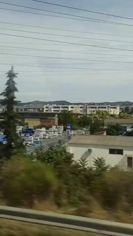 The video shows a view from a moving vehicle, likely a train or bus, passing by a street scene. On the left, there are trees and bushes. In the center and right, there are several buildings, some of which appear to be commercial establishments or warehouses. One building has large windows and signage in Greek that includes "ΚΑΤΑΣΤΗΜΑ" (store) and "ΞΕΝΟΔΟΧΕΙΟ" (hotel). Another building has a prominent red sign with Greek lettering that reads "ΥΙΟΙ Σ. ΦΡΑΝΤΖΗ Α.Ε." (Sons of S. Frantzis S.A.). Piles of materials, possibly stone or concrete slabs, are visible in front of some of the buildings. Overhead power lines are visible against a cloudy sky. The overall impression is a view of an industrial or commercial area in Greece, seen in passing.