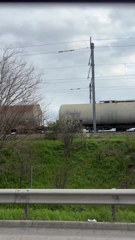 The video shows a view from inside a vehicle, looking out the side window at a moving freight train. The train consists of tanker cars, some appearing weathered with rust-like discoloration. The landscape adjacent to the train tracks features green vegetation and some bare trees. Overhead power lines are visible against a cloudy sky. A guardrail partially obstructs the lower portion of the view.