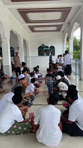 The video shows a group of young boys sitting on the floor in what appears to be an outdoor covered area, possibly a mosque courtyard or veranda. They are all dressed in traditional Islamic attire, with many wearing peci hats. Some of the boys are holding cups and drinking, while others are engaged in conversation. In the background, several adults are also present, some observing the children and others interacting with them. The environment is clean and well-maintained, with white walls and columns visible. The overall scene suggests a gathering or event involving the local community's children.