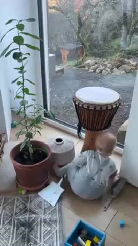 A baby wearing a light-grey onesie with suspenders is near a window in a well-lit room. The baby is on the floor, lying on its side with its head near a white wall and feet pointing towards a potted plant. The plant is green and placed on a decorative rug. Adjacent to the baby is a small, white shovel and a grey brush. Beside the baby and the plant is a small, round, grey decorative vase. Behind the baby, closer to the window, is a djembe drum on a wooden stand. The window looks out onto a yard with trees and some rocks. A blue container with colorful toys is in the bottom right corner of the frame. The floor is wooden and has a light-brown finish.