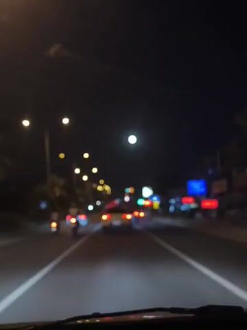 The video captures a first-person perspective of driving at night on a road. The scene is dark with streetlights illuminating the road and the surrounding environment. Several cars are visible on the road, some with their headlights on, creating streaks of light. The moon is also visible in the sky as a bright orb. The view includes the dashboard of the vehicle, suggesting the camera is mounted inside. There are no people clearly visible, but the presence of vehicles and streetlights indicates a populated or developed area. The overall atmosphere is one of night travel, with the blurred nature of the footage adding to a sense of motion.