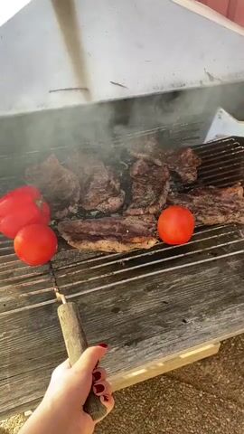 A person's hand with red nail polish is holding a barbecue fork with a wooden handle. The fork is positioned over a metal grill that contains several steaks, a red bell pepper, and two red tomatoes. The steaks are on the grill, and smoke is rising from them, indicating they are cooking. The grill is placed on a wooden surface, which appears to be part of an outdoor barbecue pit. The overall scene suggests food is being prepared on a grill outdoors.
