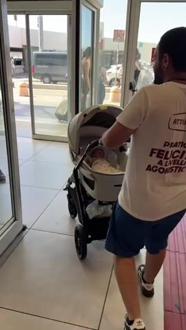 A man in a white t-shirt and blue shorts is pushing a baby stroller through an automatic sliding glass door. The t-shirt has a burgundy print that reads "ATTENZIONE PRATICO FELICITA A LIVELLI AGONISTICI!!". Another person in a white t-shirt is partially visible to the left. The man stops the stroller and looks down at the baby inside. A woman with red hair walks by carrying a white suitcase on wheels. Outside the glass doors, there is a street with people, cars, and a building with the text "Nuova Sport Car" visible. The scene appears to be at the entrance of an airport or similar building.