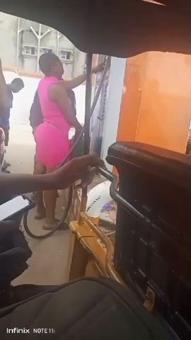 The video shows the view from inside a three-wheeled vehicle, likely a type of auto rickshaw, at what appears to be a gas station. The perspective is from the driver's seat. Several people are visible outside the vehicle. A woman in a bright pink dress is operating a gas pump. Other people are standing nearby. One person is looking at a mobile phone. The background includes a light-colored building. The quality of the video is somewhat grainy.