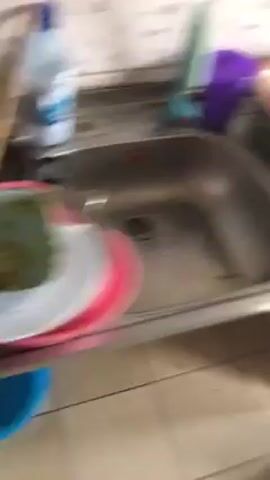 The video shows a kitchen counter cluttered with various plastic containers, some with lids, stacked on top of each other. There are also plates and a black rectangular container. A yellow container has a black utensil inside. The counter appears to be stainless steel, and the wall behind it is tiled with a speckled pattern. Part of a gray garbage can is visible on the right. The floor is tiled in a light color.