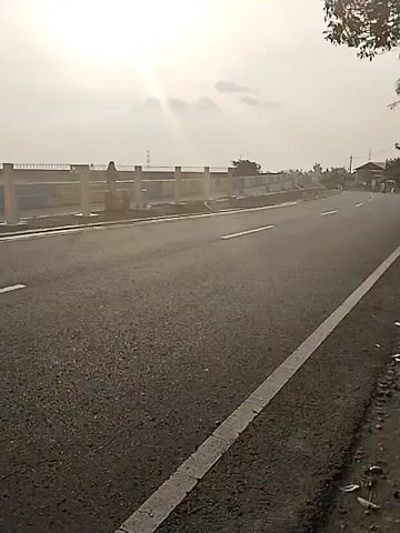 The video shows a person walking along the railing of a bridge beside an asphalt road. The person is walking from left to right across the screen, with their back to the camera, and the sunlight is visible in the sky. The road has white lane markings. Powerlines can be seen in the distance.