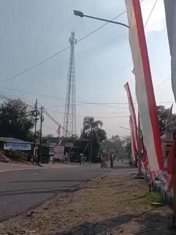 The video shows a street scene with a tall telecommunications tower in the background. On the right side of the road, several red and white flags are displayed vertically. A gray pickup truck is driving on the left side of the road. In the distance, there are some buildings and trees. The sky is clear and sunny. The video appears to be taken in a rural or suburban area.