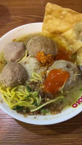 This close-up, eye-level shot focuses on a steaming bowl of Indonesian meatballs, known as "bakso," served on a warm, wooden table. The dish is a vibrant ensemble of plump, grayish-brown meatballs submerged in a clear, savory broth. Intertwined within the broth are delicate white rice noodles and bright yellow egg noodles, adding textural contrast. Garnishes include chopped green onions scattered across the surface and a dollop of spicy red chili paste.

In the upper right corner of the bowl, a piece of crispy fried wonton skin adds another layer of visual interest and a hint of crunch. The lighting suggests an indoor setting, likely a restaurant given the style of the bowl and the wooden table. The overall impression is one of a comforting and delicious meal being prepared for consumption.

The activity captured is the final preparation of the dish. In the first two seconds, a green condiment dispenser is seen hovering over the bowl, and in the third second, a dark liquid, likely soy sauce or a similar savory sauce, is being drizzled onto the meatballs. This suggests the dish is being seasoned to the diner's preference just before being served. The image captures a moment of anticipation and culinary enjoyment. The visible text on the rim of the bowl, though partially obscured, appears to be a logo or brand name in red and white. The time of day is not explicitly evident, but the warm lighting and presentation suggest it could be any time of day when a hearty meal is desired.