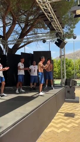 A group of young men are on a raised platform outdoors. The platform is made of wood and has a metal truss structure above it with speakers and lights. In the background, there are black screens, trees, and a building. The men are dressed casually, mostly in blue shorts and white shirts. Some of the men are shirtless. One man in the center is holding a microphone and appears to be speaking or singing. Another man is holding a phone. The men are interacting with each other and seem to be performing or participating in an event. The lighting suggests it is daytime.