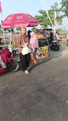 A woman wearing a hijab and a brown plaid shirt walks towards the camera carrying a white plastic bag in her right hand and a beige handbag in her left. She is wearing black pants and white platform sandals. Behind her, a red motorcycle is parked. To her right, a red umbrella with the Torabika logo is set up over a food stall. Several people are visible at the stall. A man in a patterned shirt rides a black motorcycle away from the stall. A young boy stands near the stall. The ground is paved. In the background, there are trees and a fence.