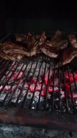 The video shows a close-up view of raw meat being grilled over glowing red coals. The meat is arranged on a metal grate, and appears to be several cuts of pork ribs or chops. The coals are emitting a bright red glow, indicating high heat. The background is dark, suggesting the grilling is taking place at night or in a covered area. The camera is positioned low, looking up at the grill from below. The video is static, with no movement from the camera or the subjects. The overall impression is one of a delicious meal being prepared.
