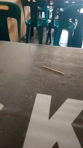The video captures a close-up of a dark-colored table, possibly made of laminate, in what appears to be an indoor setting with plastic chairs in the background. The table surface is speckled with small debris, and a single wooden toothpick lies diagonally across it. The background shows multiple teal-colored plastic chairs lined up, with a light-colored wall to the left. The lighting is somewhat dim, and the focus is primarily on the table's surface.