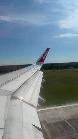 The video shows a view from an airplane window, capturing the wing of an Air Asia plane against a backdrop of a clear blue sky with scattered clouds. The plane appears to be taxiing or preparing for takeoff on a runway, indicated by the marked concrete surface visible beneath the wing. Beyond the runway, there's a green, grassy area with rows of palm trees in the distance. The Air Asia logo is prominently displayed on the red and white tail fin of the plane.