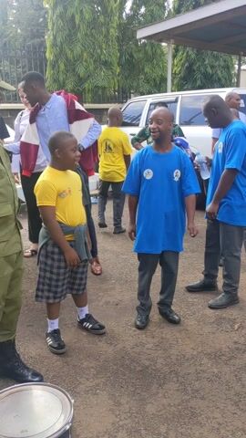 The video shows a group of people outdoors, with two of them, likely young adults, in the foreground. One is wearing a blue t-shirt and the other, a yellow t-shirt with plaid shorts. They are smiling and appear to be interacting with a man in a green military-style uniform. In the background, there is a group of other people, including some wearing similar yellow or blue shirts, a few adults, and a white SUV. A drum sits on the ground to the left. The environment suggests a paved area possibly near a building, with trees in the background. There appear to be friendly interactions occurring between the people.