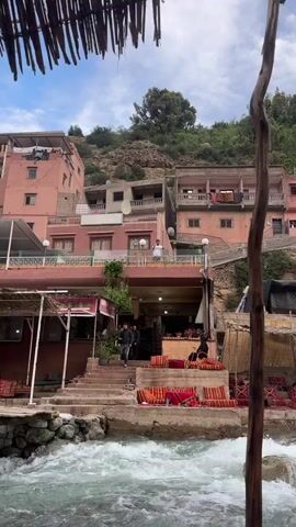 The camera films from across a river, focusing on a man standing on the second-story balcony of a pink building, waving and making heart shapes with his hands toward the camera. The river runs rapidly, lined with rocks. The pink building has a restaurant on the ground level with outdoor seating. Other people can be seen walking on steps and sitting at tables. Behind the buildings, there is a steep, green, rocky hill. The sky is overcast.