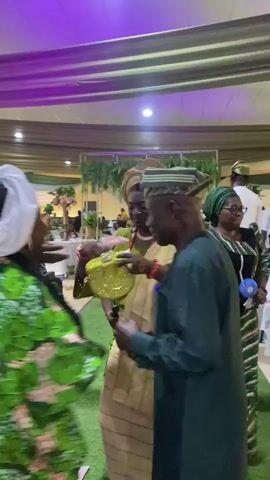 The video shows a gathering of people, seemingly at a celebration or event. Several individuals are dressed in traditional attire, with some wearing colorful, patterned fabrics and head coverings. Others are in more contemporary clothing. The setting appears to be an indoor event space decorated with green and white draperies, indicating a possible Nigerian theme. People are interacting with one another, exchanging greetings, and some are smiling and laughing. The atmosphere seems festive and social. There are other details such as a glimpse of floral arrangements and a ceiling fan.