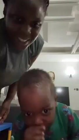 A woman and baby are in a room. The woman is standing behind the baby who is on the floor. The woman is smiling at the camera and talking. The baby is smiling and making sounds. The room has a white ceiling with inset lighting. A TV and picture frame are on the wall. A blue table is in the background. The woman is wearing a gray sleeveless shirt and black shorts. The baby is wearing a green patterned shirt.
