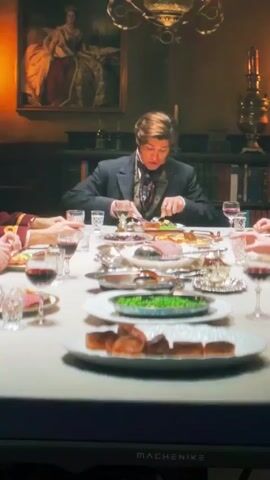 The video shows a formal dinner scene. A man in a dark suit and cravat is seated at a large table, cutting into a plate of food. There are other people at the table, mostly their hands and arms are visible. The table is laden with platters of food, including what appears to be roasted meat and vegetables. There are wine glasses filled with red wine. In the background, a large portrait of a woman in a regal gown hangs on the wall, and bookshelves filled with books are visible. Another clip shows a close-up of a man with dark curly hair and a mustache, wearing a red military-style uniform with gold braid and buttons. The overall setting suggests a historical period.