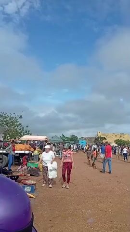 The video shows an outdoor market scene with many people walking around and browsing stalls. The ground is dirt, and there are various tents and umbrellas providing shade. Some people are carrying bags, suggesting they are shopping. In the background, there are buildings and vehicles, including cars and trucks. The sky is blue with some clouds. The overall atmosphere appears to be that of a busy marketplace on a sunny day.