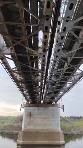 The camera faces upwards from below a large metal truss bridge. The bridge has two parallel spans supported by a thick concrete pillar rising from the water below. The metal structure of the bridge is heavily riveted and shows signs of rust in places. The sky is overcast and a low, wooded shoreline is visible in the distance. No people or other objects appear in the video. The camera slowly pans upward, tracing the structure of the bridge.