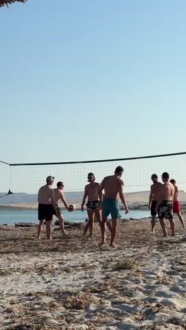 A group of eight young men are playing volleyball on a sandy beach near a body of water. A volleyball net is set up in the center of the frame. The sky is clear and blue. The men are all wearing swim trunks. They are in various stages of playing the game, with some reaching for the ball and others watching. The beach is covered in sand and some dry vegetation. The background shows a body of water and distant hills.