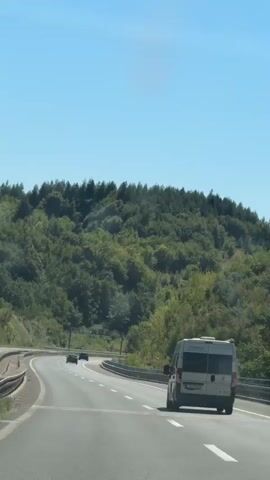 The video shows a view from a vehicle traveling on a highway. The highway has multiple lanes and is bordered by guardrails. On the right side of the road, a white van with tinted windows is visible, with a portion of its license plate visible. In the distance on the highway, two dark-colored cars are driving ahead of the camera. The landscape outside the highway consists of a densely wooded hill with green trees under a clear blue sky with a bright sun. The road appears to be curving to the left.