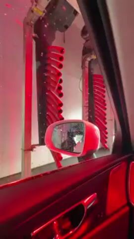 The view from inside a red car shows it moving through a car wash tunnel. The car’s side mirror is red, and two arrays of red nozzles on dark hoses hang from the ceiling.  One array has roughly twenty nozzles and the other has roughly ten nozzles. The car wash tunnel is lit by red lighting. Water spray is visible in the tunnel.