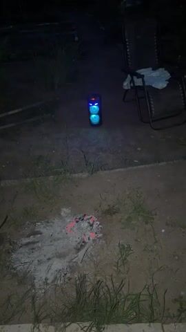 This short video, likely recorded in Nezhinka, Russia, captures a nighttime scene in an outdoor setting. The primary object of focus is a portable speaker with colorful, pulsating LED lights. The speaker is positioned on the ground, illuminating the immediate area with changing red, green, and blue hues.

In the foreground, there is evidence of a recent fire. Embers glow red within a pile of ash and debris, suggesting a campfire or bonfire has recently been extinguished. Surrounding the ash pile is dry, unkempt grass and dirt.

Further back, to the right of the speaker, a dark metal lawn chair with a light-colored fabric seat is visible. The chair appears empty. In the background, a dark fence and some greenery can be seen, indicating a yard or garden area.

The time of day is clearly night, with darkness enveloping the scene except for the artificial light from the speaker and the dying embers. The overall mood is somewhat subdued and relaxed, possibly hinting at a quiet evening outdoors after a fire. There is no visible text in the media.