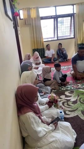 A group of people are gathered indoors. In the foreground, several women in hijabs are seated on the floor. One woman in a pink hijab is holding a bouquet of flowers and a smartphone. Beside her, another woman in a white hijab is also holding a smartphone. To their right, a young boy is seated on the floor. Further back, a group of men are seated on the floor, some wearing skullcaps. The room has yellow walls and a large window with yellow curtains. A patterned rug covers the floor. Two framed documents are hanging on the wall. The scene appears to be a gathering, possibly a religious ceremony or a family event.