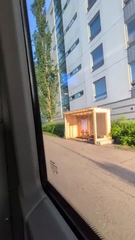 The video is filmed from inside a vehicle, looking out the passenger window. The vehicle is moving along a paved road next to a sidewalk. To the right of the road, there are several apartment buildings, tall and modern with many windows. Adjacent to the road and sidewalk, there are green bushes and trees. In the foreground, a partially constructed wooden bus shelter is visible. Further down the road, a standard bus stop sign with route information is present, accompanied by yellow traffic cones. The sky is clear and blue, indicating it is daytime with good weather. Sunlight reflects off the buildings and creates shadows on the road.