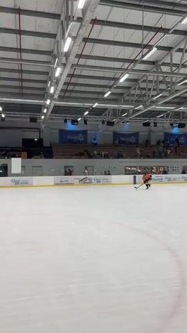 A hockey game is being played on an indoor ice rink. The rink has glass boards around it, and spectators can be seen in the bleachers in the background. A scoreboard indicates the time remaining in the period and the score. One player in an orange jersey is skating towards the goal with the puck. Several players in green jerseys are on defense, with one player in goal. A referee in black and white stripes is also on the ice. The ceiling of the arena is visible, with exposed beams and lighting fixtures.