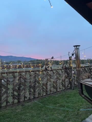 The video presents a serene outdoor scene during twilight, likely dusk or dawn, in Tyulyuk, Russia. The sky displays soft hues of pink blending into light blue above a distant range of mountains. In the foreground, a decorative wooden fence with a distinctive geometric pattern runs horizontally across the frame. String lights with warm yellow glows are draped along the top of the fence, providing artificial illumination.

Several black birds, resembling crows or ravens, are perched along the fence and on a wooden post to the left. A small, dark wooden birdhouse or feeder is also visible on this post. Behind the fence, green grass stretches towards the background, where a few modest buildings, including two white, dome-shaped structures and some wooden cabins, can be seen. Partially visible behind the fence and string lights is a wooden sign with Cyrillic text, where the word "ГОРНЫЕ" (GORNЫE), meaning "Mountain," is discernible. On the far right, a section of a metal chimney or drainpipe is visible. The scene evokes a tranquil and peaceful atmosphere.