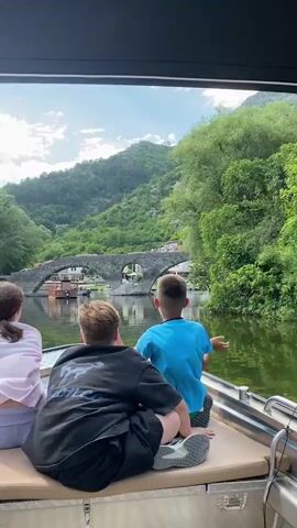 The video shows three children on a boat looking out at a body of water and a stone bridge in the distance. The child in the foreground is wearing a black shirt and shorts, and is partially obscured by the camera's perspective. A second child in a blue shirt and green shorts sits to the right, facing forward. A third child, mostly obscured by the foreground child, is wearing a pink shirt and white shorts. The boat appears to be moving slowly, and the water is calm. In the background, a large stone bridge spans the water, with buildings and lush green vegetation visible on the hillsides beyond. The sky is partly cloudy.