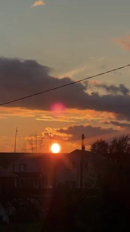 A vertical video begins with a sunset over a rural landscape. The sun is a bright orb near the horizon, casting warm orange and pink hues across the sky. Clouds are scattered, some dark and others illuminated by the setting sun. In the foreground, the rooftops of houses and trees are visible, silhouetted against the vibrant sky. Electrical wires and a utility pole stretch across the frame, indicating a settled area. As the video progresses, the sun dips lower, and the colors in the sky intensify. The camera pans slightly, revealing more of the surrounding greenery and the tops of trees. The video ends with the sky filled with soft, pastel colors and fluffy clouds.
