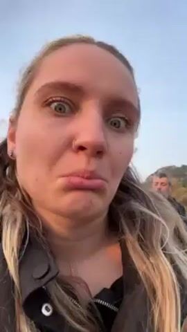 A woman with long blonde hair, fair skin, and a nose ring is centered in the frame, making a face with wide eyes and pursed lips. She wears a black jacket. Behind her, slightly out of focus, stands a man with short grey hair. The background consists of trees and a hazy sky. The video appears to be a selfie or close-up recording taken outdoors.