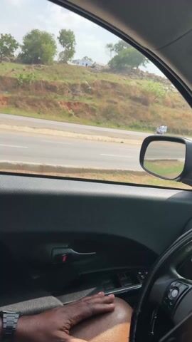 From the perspective of the driver of a car, a wrecked dump truck is visible on the side of the road. Other cars and motorcycles pass by. The road is bordered by a grassy hill with scattered trees and a building.  The driver's hand and arm, adorned with a watch, are visible in the foreground. Music plays.