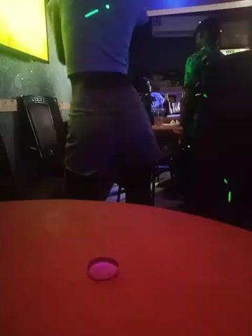 In a dimly lit indoor space, possibly a bar or club, a person dances facing away from the camera. The dancer wears a white t-shirt and shorts. Green and pink laser lights dot the scene, highlighting the dancer and other people and objects. In the background, tables and chairs are visible. A bottle cap sits on the orange table in the foreground.