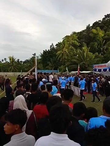 This video captures a lively outdoor event in Keude Trienggadeng, Indonesia. The scene is bustling with a large crowd of people, many of whom are wearing bright blue t-shirts with white lettering, suggesting they might be part of a group or organization participating in the event. The crowd is gathered in an open grassy area, with a backdrop of lush green trees and a hillside in the distance. A white tent with blue trim is set up, likely serving as a stage or information booth, and it displays prominent text.

The event appears to be a public gathering, possibly a festival, celebration, or official ceremony. The presence of uniformed individuals, including what appears to be police officers in dark uniforms, indicates a potentially official capacity for the event. The mood seems festive and engaging, with people of all ages milling about and observing the proceedings. Some individuals are gathered closely around a central area where a few people are standing, possibly giving speeches or performing.

The time of day appears to be late afternoon or early evening, given the soft lighting and the sky's appearance. The weather seems pleasant, suitable for an outdoor event. The overall impression is one of community and shared activity, with a strong sense of local participation.

Visible text on the tent includes "HARI BATANGKARA" and "POLRI RESOR PIDIE JAYA" along with a date that reads "41 TAHUN 2020." This text provides strong clues about the event, suggesting it is related to the anniversary of the Pidie Jaya Police Resort, perhaps a "Hari Bhayangkara" (National Police Day) celebration. The numerical "41" and the year "2020" specify the anniversary number and the year the event is commemorating.