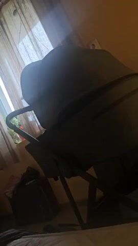The video shows a gray stroller in a room with a window. The stroller has a canopy and a dark frame. The room has orange walls and a white bedspread. There is a bag on the floor next to the stroller. Light streams in through the window. The camera is handheld and shaky, causing the stroller to move in the frame.
