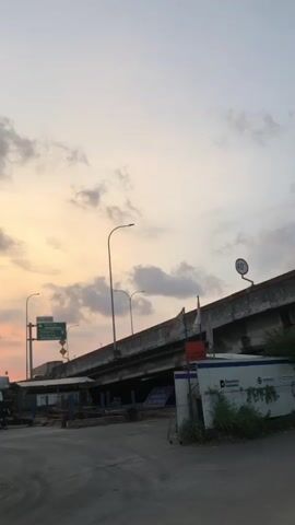 The video captures an outdoor scene under a cloudy sky with a soft, diffused light suggesting either sunrise or sunset. The primary focus is an elevated concrete structure, possibly a highway overpass or bridge, which occupies the upper right portion of the frame. The overpass has visible signs and lampposts attached to its sides. Below the overpass, there appears to be some construction or maintenance activity, with what looks like metal beams or scaffolding.

In the foreground, there is a paved area, likely a road or a parking lot, with some vegetation growing along the edges. To the right, a white portable building or container is visible, bearing some text and logos, one of which appears to be "Departure..." suggesting it might be related to transportation or logistics. A red and orange sign is also attached to this structure. No people are visible in the video. The overall environment appears to be an urban or semi-urban setting, possibly near a transportation hub. The lighting and clouds create a calm, somewhat muted atmosphere.