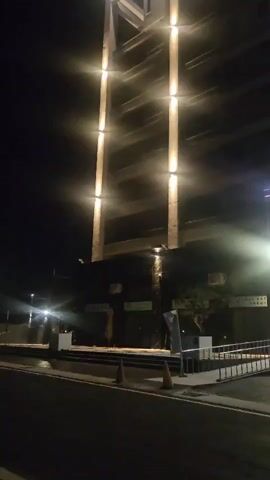 A video shows the exterior of a modern building at night. The building is illuminated by vertical lights that run up its facade. The entrance area features a ramp, a set of stairs, and a large glass facade with decorative elements. The street in front of the building is wet, reflecting the lights. The video appears to be filmed from a low angle, across the street from the building. There are traffic cones on the sidewalk. The overall impression is of a contemporary architectural structure bathed in artificial light after dark.