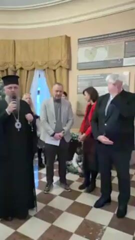 Here is a detailed description of the video:

The video shows a small group of people gathered indoors in what appears to be a museum or public building, indicated by the tiled floor, high ceilings, and informational displays on the walls. The main focus is on three men standing relatively close together. On the left, a man dressed in a traditional Orthodox Christian priest's garb, complete with a tall hat and a cross hanging from his neck, is speaking into a microphone. In the center stands a man in a casual blazer, while a man in a dark suit stands to the right. The man in the suit appears to be wiping his eye. A few other people are visible in the background, including a woman in a red jacket. In front of the man in the blazer is a box decorated with foliage, and what looks like a person inside the box. The priest is gesturing while speaking, and the others are listening. The overall atmosphere is formal but intimate.