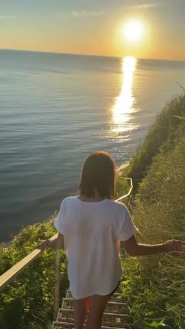 A young woman with dark hair is walking down a metal staircase that is covered in green foliage. She is wearing a white t-shirt and dark shorts. The staircase is next to a steep, grassy incline that overlooks a calm body of water. The sun is setting, casting a golden glow across the water and the sky. The woman turns to look back at the camera with a smile.