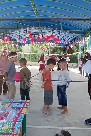 The video shows a group of young boys participating in a game under a blue canopy. The boys are standing in a line, with their legs tied together. They are all barefoot. Some boys are holding balloons. The ground is paved and there is a table with cups and plastic bags on the left side of the frame. The background shows trees and buildings. The canopy is decorated with red and white balloons and flags. A man in a blue shirt and black pants is standing on the right side of the frame, possibly supervising the game.